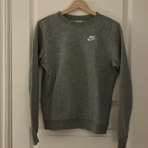 Grå sweatshirt från Nike - Säljer en stilren grå sweatshirt från Nike med rund hals och långa ärmar. Tröjan har en vit Nike-logga på bröstet och är perfekt för en avslappnad stil. Passar bra till både jeans och träningsbyxor.