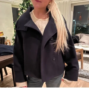 Svart kappa med knappar - Snygg svart kappa med hög krage och knappar framtill. Jackan har en stilren design med långa ärmar och en rak passform. Perfekt för kyligare dagar och ger en elegant look.