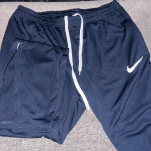 Svarta träningssbyxor från Nike - Snygga svarta träningsshorts från Nike med vit dragsko i midjan och en dragkedjeficka på sidan. Perfekta för sport och träning. Nike-loggan syns tydligt på benet.