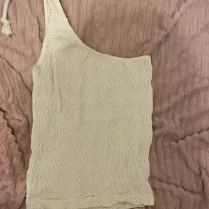  oneshoulder topp - Säljer en stilren vanilj vit/vit beige oneshoulder topp med textur. Perfekt för en trendig look. Toppen har en unik design med en axel och är i ett mjukt material.