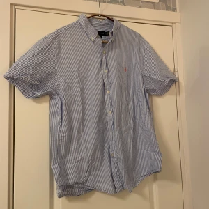 Blå och vit randig skjorta från Ralph Lauren - Säljer en klassisk blå och vit randig skjorta från Ralph Lauren. Skjortan har korta ärmar och knappar framtill. Perfekt för en stilren look.