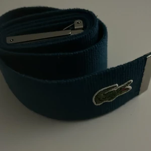 Blått bälte från Lacoste - Snyggt blått bälte från Lacoste med en silverfärgad spänne. Bältet har den klassiska krokodilloggan och texten 'Life is a beautiful sport' ingraverad på spännet. Perfekt för att ge en sportig touch till din outfit. Helt ny, nypris 500 kr mitt pris = 199