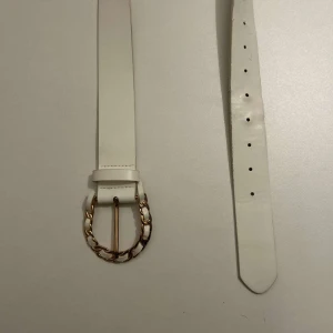 Vitt bälte med gulddetaljer - Snyggt vitt bälte med en unik guldfärgad spänne som har en kedjedesign. Perfekt för att ge en stilren touch till din outfit. Bältet har flera hål för justerbar passform.
