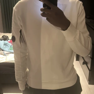 Vit sweatshirt från C.P. Company - Säljer en stilren vit sweatshirt från C.P. Company. Tröjan har långa ärmar och en rund halsringning. Den är prydd med en rund detalj på ärmen. Bara använt 3 gånger 