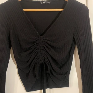 Svart ribbad tröja med knyt - Snygg svart ribbad tröja i storlek XS. Tröjan har en v-ringad design med knytning framtill som ger en fin rynkad Loke. 