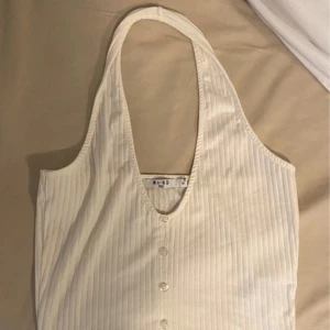 Vit ribbad halterneck topp från NA-KD - Säljer en stilren vit ribbad halterneck topp från NA-KD. Toppen har knappar framtill och en snygg v-ringning. Perfekt för varma dagar eller som en del av en lager-på-lager look.
