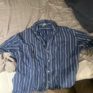 Blå och vit randig skjorta från Acne Studios - Snygg blå skjorta med vita ränder från Calvin Klein. Skjortan har en klassisk krage och knappar framtill. Perfekt för en stilren look. Strl 48 oversized och sitter som L men kan funka M beroende vad man själv tycker. Knappt använd och nypris 3800 kr