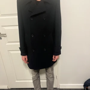 Svart skepparkavaj från Dressmann - En mörkblå ullrock från Dressmann i gott skick. Storlek 52/M, passar perfekt för någon som är 180 cm lång. Kavajen har en klassisk dubbelknäppt design med knappar framtill och långa ärmar. Perfekt för en elegant look.
