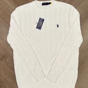Vit stickad tröja från Ralph Lauren - Säljer en stilren vit stickad tröja från Ralph Lauren med det klassiska broderade logotypen på bröstet. Den är helt ny och prislapp ingår. Tröjan har en kabelstickad design och långa ärmar med ribbade muddar. Perfekt för en elegant och avslappnad look.