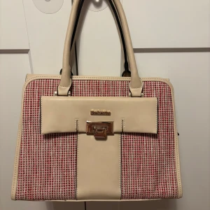 Beige och röd handväska från Privata - Snygg handväska i beige och rött från Privata. Väskan har ett rutigt mönster på framsidan och en praktisk dragkedja på baksidan. Insidan är fodrad med ett randigt mönster och har flera fack för enkel organisering. Perfekt väska för hösten med mycket plats i.