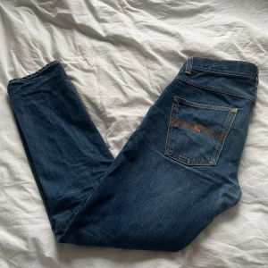 Mörkblå jeans från Nudie Jeans - Snygga mörkblå jeans från Nudie Jeans med klassisk femficksdesign och orange sömmar. Perfekta för en avslappnad stil. De har en normal passform och är tillverkade i slitstarkt denim. Dom har hål i högra fram fickan men syns inte utanpå byxorna
