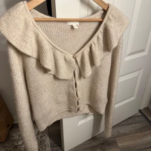 Beige kofta med volangdetalj - Säljer en mysig beige kofta med volangdetalj längs halsringningen. Koftan har långa ärmar och knappar framtill. Perfekt för en stilren och bekväm look.