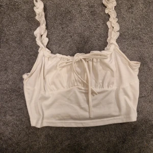 Vit linnetopp med volangdetaljer - Söt vit linnetopp med volangprydda axelband och knytning framtill. Perfekt för varma sommardagar eller som ett lager under en kofta. Toppen har en mjuk och bekväm passform.