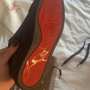 Svarta sneakers med nitar från Christian Louboutin - Snygga svarta sneakers från Christian Louboutin med ikonisk röd sula och nitar på tån. Skorna har snörning och är tillverkade i mocka. Perfekta för att ge en edgy touch till din stil.