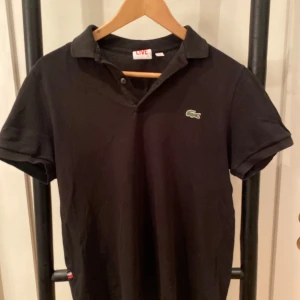 Svart pikétröja från Lacoste - Snygg svart pikétröja från Lacoste med klassisk krage och korta ärmar. Liten i storleken M.