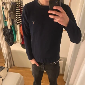 Mörkblå tröja från Gant - Säljer en stilren mörkblå tröja från Gant med broderad logga på bröstet. Tröjan har långa ärmar och ribbade muddar vid ärmslut och nederkant. Perfekt för en avslappnad men snygg look.