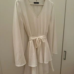 Vit klänning från rut & circle - Elegant vit klänning med v-ringning och knytband i midjan. Klänningen har långa ärmar och en flödande design som ger en fin siluett. Perfekt för en stilren look. Inga tecken på användning. Går att använda med eller utan band, medföljer 