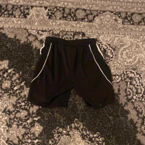 Svarta shorts med vita detaljer - Säljer ett par svarta shorts med vita ränder längs sidorna. Perfekta för träning eller en avslappnad dag. De har en elastisk midja för extra komfort.