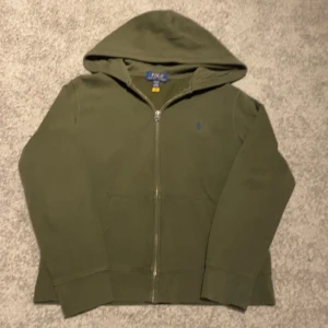Olivgrön hoodie från Ralph Lauren - Säljer en olivgrön hoodie från Ralph Lauren med dragkedja och huva. Tröjan har långa ärmar och en liten logga på bröstet. Perfekt för en avslappnad stil.      Tröjan är storleken 14/16 vilket är en xs