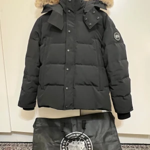  Canada Goose Wyndham - Säljer en svart dunjacka från Canada Goose med avtagbar huva och pälsdetalj. Jackan har en quiltad design med knappar och dragkedja framtill. Perfekt för kalla vinterdagar.