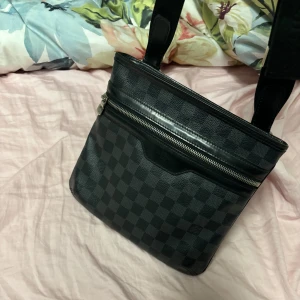 Svart axelväska från Louis Vuitton - Snygg svart axelväska från Louis Vuitton med klassiskt rutmönster. Väskan har en praktisk dragkedja framtill och justerbar axelrem. Perfekt för att bära dina dagliga nödvändigheter med stil. Använd ca 2-3 gånger sedan har den bara hängt som prydnad. Riktigt fint väska, äkta såklart!
