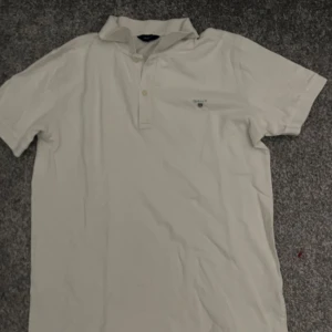 Vit kortärmad pikétröja från GANT - Säljer en stilren vit pikétröja från GANT med korta ärmar och klassisk krage. Tröjan har knappar framtill och en liten broderad logotyp på bröstet. Perfekt för en avslappnad men ändå snygg look.