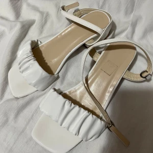 Vita sandaler med klack - Snygga vita sandaler med klack och rynkad detalj framtill. De har en justerbar rem runt ankeln och en fyrkantig tå. Perfekta för sommarens alla tillfällen.
