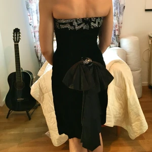 Vintage cocktailklänning - Elegant svart axelbandslös cocktailklänning med pärldetaljer längs överkanten. Klänningen har en stor rosett baktill och är perfekt för festliga tillfällen så som jul och nyår. Ganska liten i storlek.