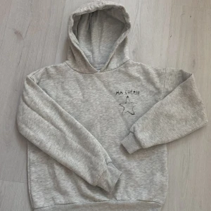 Grå hoodie med tryck 'Ma Chérie' - Säljer en mysig grå hoodie med texten 'Ma Chérie' och en stjärna tryckt på bröstet. Tröjan har en klassisk huva och ribbade muddar vid ärmslut och nederkant. Perfekt till vardags 