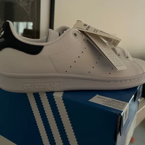 Vita Adidas Stan Smith sneakers - Klassiska vita helt oanvända Adidas Stan Smith sneakers med marinblå detalj på hälen. Perfekta för en stilren look. Skorna har snörning och är tillverkade i syntetmaterial.
