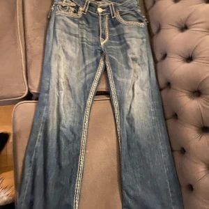 Blå jeans från True Religion - Säljer ett par snygga blå jeans från True Religion med kontrastsömmar och knappar. De har en klassisk femficksdesign och en bootcut-stil som ger en avslappnad look. Perfekta för dig som vill ha en tidlös stil med en modern twist.