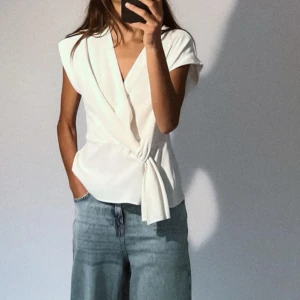 Vit omlottblus från Zara - Elegant vit omlottblus från Zara med knytning i sidan. Blusen har en v-ringad design och är ärmlös, vilket gör den perfekt! 
