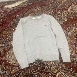 Grå sweatshirt från Lacoste - Säljer en stilren grå sweatshirt från Lacoste med det klassiska krokodilmärket på bröstet. Tröjan har långa ärmar och en rund halsringning. Perfekt för en avslappnad look.