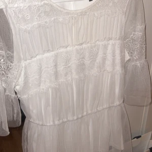 Vit spets blus  - Charmig vit blus  med spetsdetaljer och transparenta ärmar. Tröjan har en rund hals och ett romantiskt utseende med volanger och spetsmönster. Perfekt för speciella tillfällen.