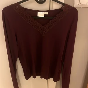Vinröd topp med spetsdetaljer från Gina Tricot - Säljer en vinröd långärmad topp från Gina Tricot med v-ringning och spetsdetaljer runt halsen. Perfekt för en stilren look.