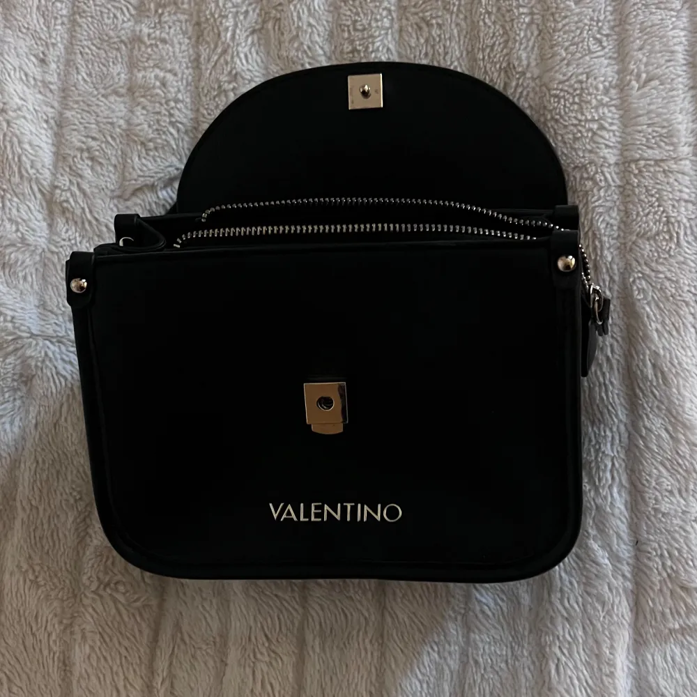 Elegant svart axelväska från Valentino med kedjedetaljer och dragkedja. Perfekt för att bära dina nödvändigheter med stil. Väskan har en stilren design med ett lock och en säker stängning.. Laukut & Käsilaukut.