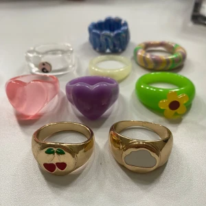 Färgglada ringar i olika former i storlek L - Säljer ett set med åtta unika ringar i olika färger och former. Inkluderar två guldiga ringar med körsbär och moln, samt färgglada plast- och akrylringar i hjärt- och blomform. Perfekt för att mixa och matcha!