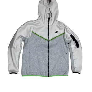 Nike tech - Säljer en grå och vit jacka från Nike med huva och dragkedja. Jackan har svarta detaljer och en grön linje som ger en sportig look. Perfekt för träning eller vardagsbruk.