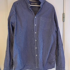 Blå och vit rutig skjorta - Snygg blå och vit rutig skjorta med klassisk krage och knappar framtill. Perfekt för en stilren look. Skjortan har långa ärmar och en bröstficka.