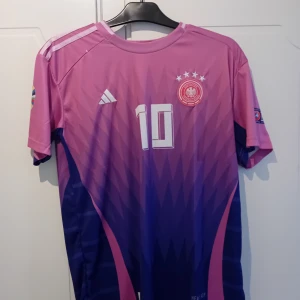 Rosa och lila fotbollströja med nummer 10 - Snygg kortärmad fotbollströja från Adidas i rosa och lila med nummer 10 på både fram- och baksidan. Tröjan har ett coolt geometriskt mönster och flera märken på ärmarna. Perfekt för fotbollsfans!