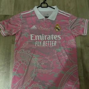 Rosa Real Madrid fotbollströja från Adidas - Snygg rosa Real Madrid fotbollströja från Adidas med ett unikt drakmönster i grått 🐉Storlek S och i 10/10 skick (aldrig använd) Perfekt för fans som vill sticka ut på läktaren eller planen. ⚽
