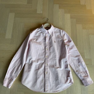Rosa och vit randig skjorta från Ralph Lauren - Snygg rosa och vit randig skjorta från Ralph Lauren med klassisk button down krage och broderad logga på bröstet. Skjortan har långa ärmar och knappar framtill. Perfekt för en stilren look! Custom fit storlek M nypris 1800kr