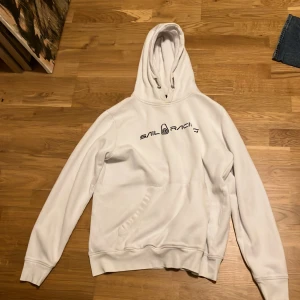 Vit hoodie från Sail Racing - Säljer en stilren vit hoodie från Sail Racing med svart logotyp på bröstet. Tröjan har en klassisk huva med dragsko och ribbade muddar vid ärmslut och nederkant. Perfekt för en avslappnad look.