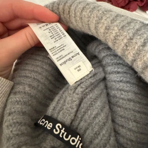 Grå stickad mössa från Acne Studios - Säljer en stilren grå stickad mössa från Acne Studios. Mössan har en ribbad design och pryds av ett ikoniskt ansiktsmärke framtill. Perfekt för att hålla värmen under kyliga dagar.