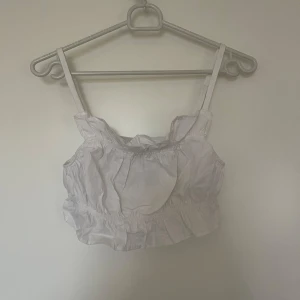Vit croppad topp från H&M - Säljer en söt vit croppad topp från H&M med justerbara axelband och volangdetaljer. Perfekt för sommardagar och kan enkelt matchas med olika outfits. Toppen är i nytt skick och har en luftig passform.