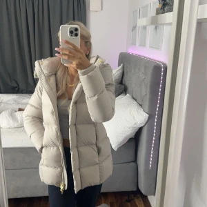 Beige pufferjacka från Rockandblue - Snygg beige pufferjacka från Rockandblue med gulddetaljer och dragkedja. Jackan har en mysig huva och är perfekt för kyliga dagar. Insidan har ett coolt mönster i guld och svart. Säljer då jag har två st. Nypris 3599