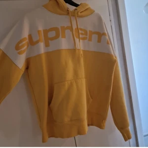 Gul och vit hoodie från Supreme Stl S - Säljer en snygg gul och vit hoodie från Supreme med stor logga över bröstet. Hoodien har en klassisk känguruficka och justerbar huva med snören. Perfekt för en avslappnad stil. Fläck som syns på bilden. Går kanske att få bort