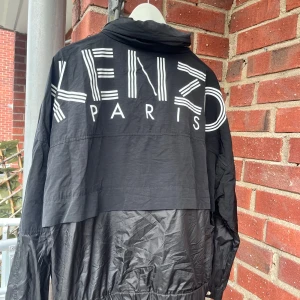 Svart vindjacka från Kenzo - Snygg svart vindjacka från Kenzo med stort tryck på ryggen. Jackan har en dragkedja i guld och en hög krage. Jackan är köpt på Kenzo i Paris för 6800:- använd en gång 