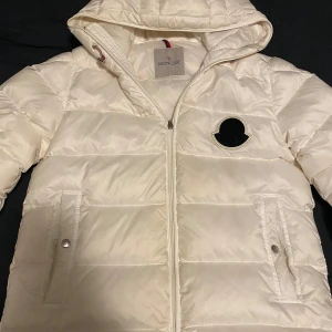 Vit dunjacka från Moncler M/S - Snygg vit dunjacka från Moncler med huva och dragkedja. Jackan har ett stilrent svart emblem på bröstet och praktiska fickor med knappar. Perfekt för kyliga dagar. Pris kan diskuteras. Storleken fungerar på både S och M 