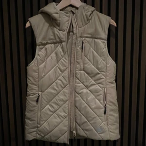 Beige quiltad väst med huva - Snygg beige quiltad väst från 157 med dragkedja och praktiska fickor. Perfekt för lager-på-lager under kyliga dagar. Västen har en bekväm huva och stilrena diagonala sömmar.